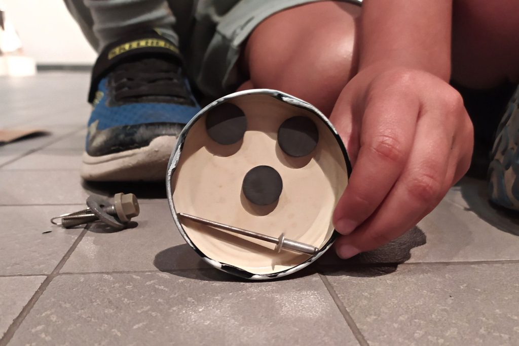 Una cara hecha con objetos metálicos e imanes apoyada en el suelo y sujeta por la mano de un niño.